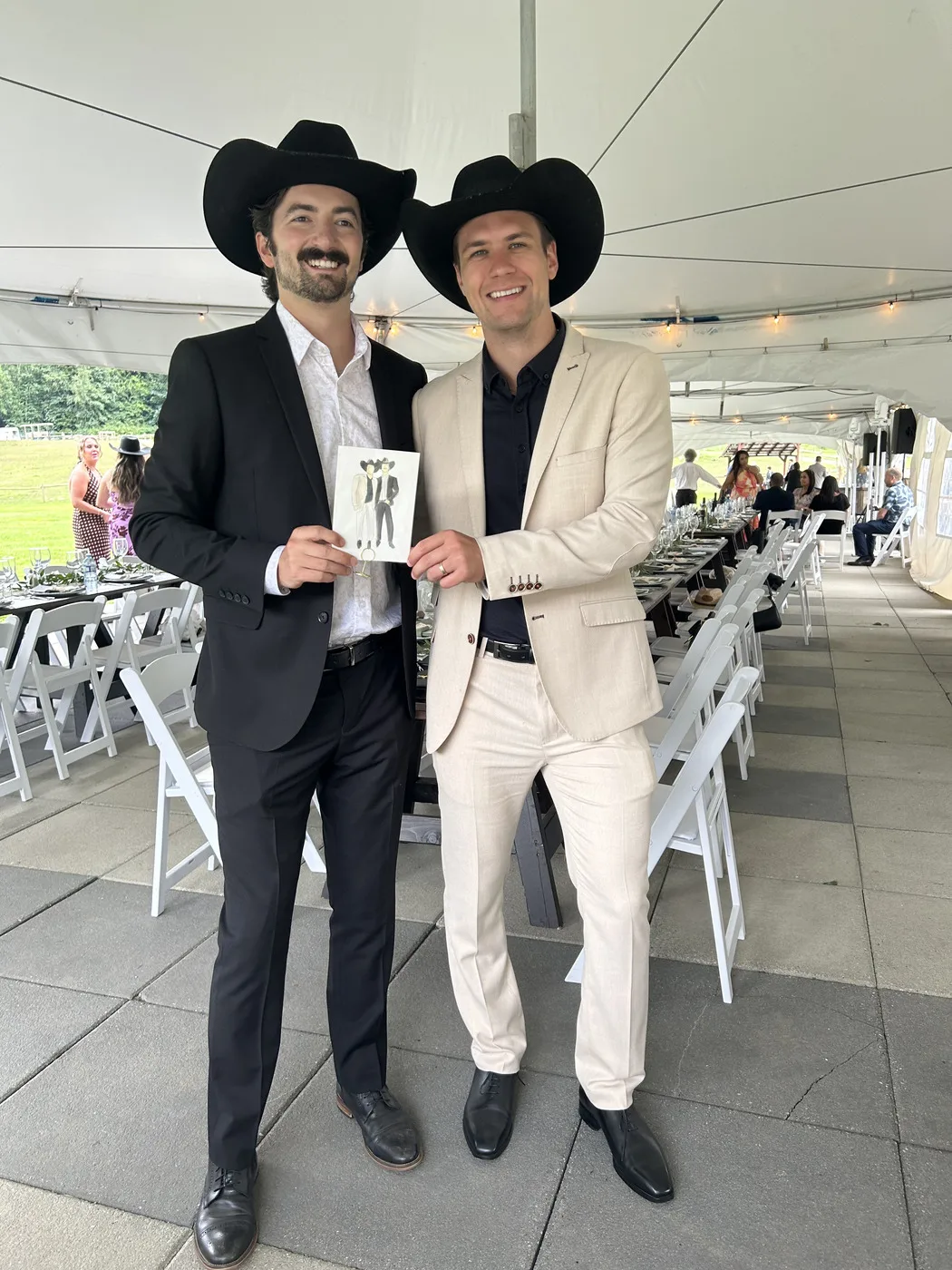 Two guests in cowboy hats holding up the watercolour illustration of themselves at a wedding reception under a white tent