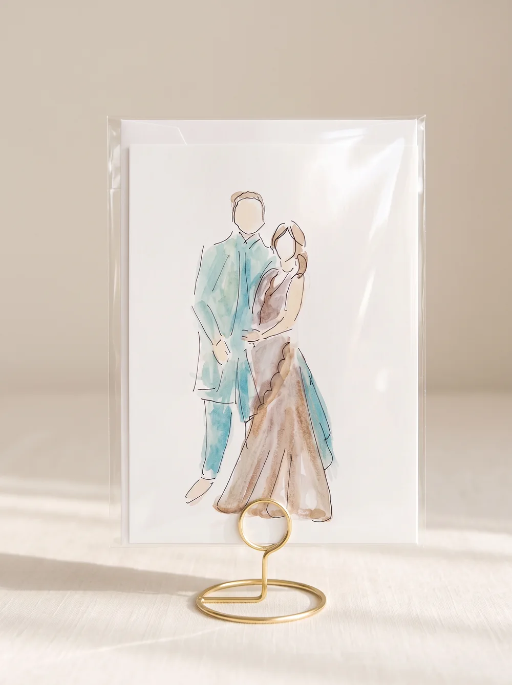 Watercolour and ink illustration of a bride and groom standing together, the groom in teal and the bride in warm taupe, inside a clear protective sleeve on a gold wire table stand