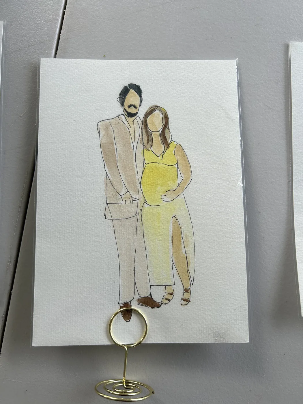 Watercolour and ink illustration of that couple, the woman in a yellow dress holding her baby bump, displayed in a gold table stand