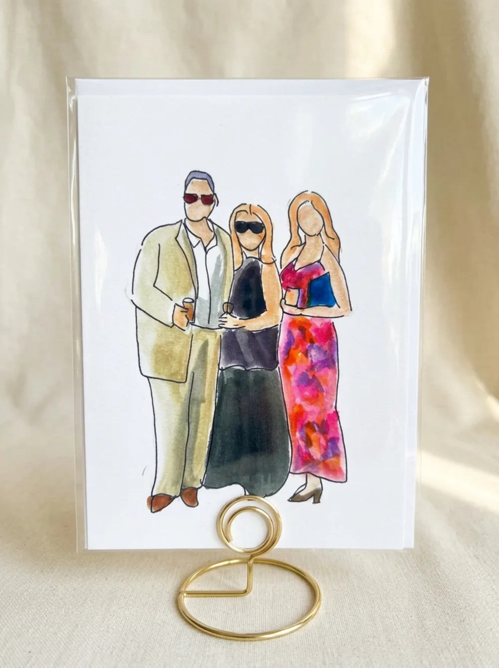 Loose watercolour illustration of three party guests, the man in sunglasses, one woman in black and one in a bright floral dress, inside a clear protective sleeve on a gold wire table stand